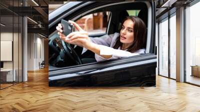 Happy young woman holding mobile phone and taking photos while driving a car. Smiling girl taking selfie picture with smart phone camera outdoors in car. Holidays and tourism concept Wall mural